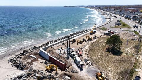 Aerial view of a coastal construction project along a shoreline road. Heavy equipment, including excavators, loaders, a crane, and concrete trucks, are working on a large seawall and shoreline protection structure. Rock armor lines the beach beside the new concrete wall, while ocean waves break along the coast. Residential homes and a roadway run parallel to the shoreline in the background under a clear blue sky.